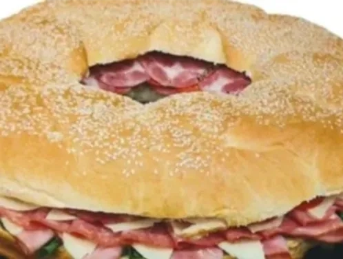A large round sandwich with a hole in the center, filled with layers of deli meat, cheese, and vegetables on a sesame seed bun.