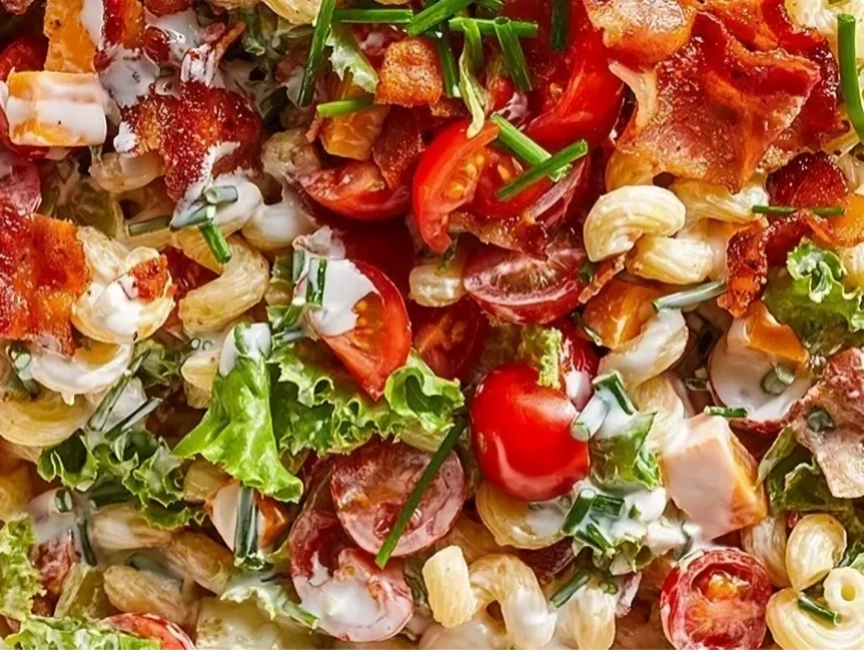 Close-up of a pasta salad with cherry tomatoes, lettuce, bacon pieces, chives, and creamy dressing.