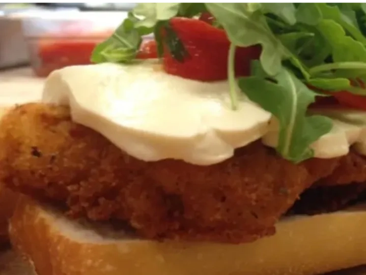 A sandwich with a breaded fried cutlet, slices of mozzarella cheese, cherry tomatoes, and fresh arugula on a slice of white bread.