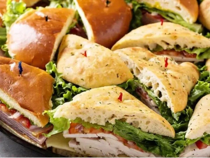 A platter of assorted sandwiches with lettuce, tomato, cheese, and deli meats on different types of bread, each sandwich held together with a colored toothpick.