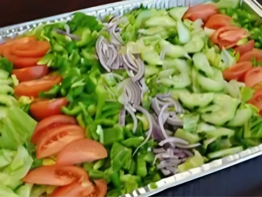 A foil tray filled with a mixed salad containing sliced tomatoes, cucumbers, green bell peppers, red onions, and lettuce.