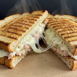 A grilled sandwich cut in half, filled with melted cheese and a creamy tuna mixture, with steam rising from the hot bread.